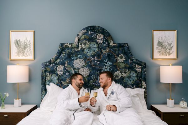 Two people in robes are sitting on a bed, toasting with champagne. The bed has a floral headboard and there are two lamps and framed art on the wall.