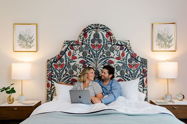 A smiling couple sits in bed with a laptop, surrounded by bedside tables and lamps, under a colorful, patterned headboard and framed art.