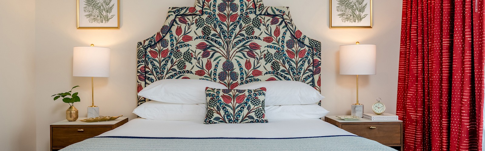 A cozy bedroom featuring a floral-patterned headboard, two bedside tables with lamps, red curtains, and framed wall art.