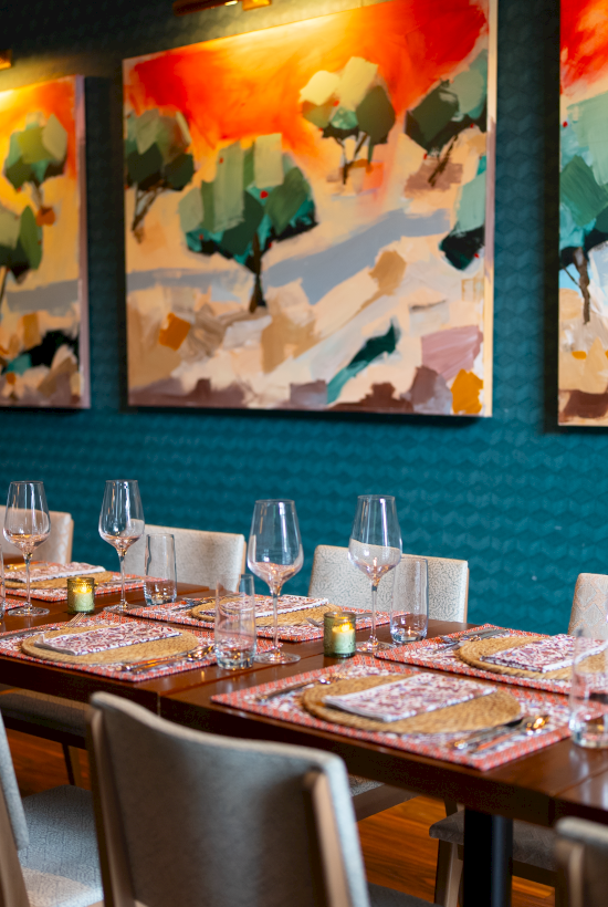 A dining area with a long table, set elegantly with glasses and tableware, under vibrant landscape paintings on a teal wall.