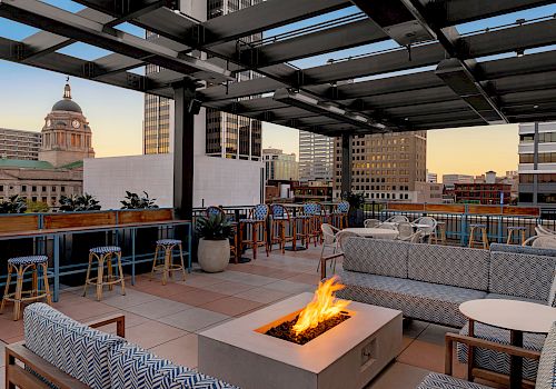 A rooftop lounge with modern furniture, a fire pit, bar seating, and a view of city buildings, including a domed structure at dusk.