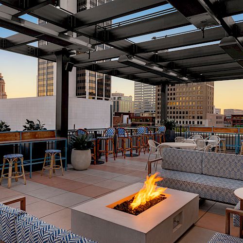 A rooftop lounge with modern furniture, a fire pit, bar seating, and a view of city buildings, including a domed structure at dusk.