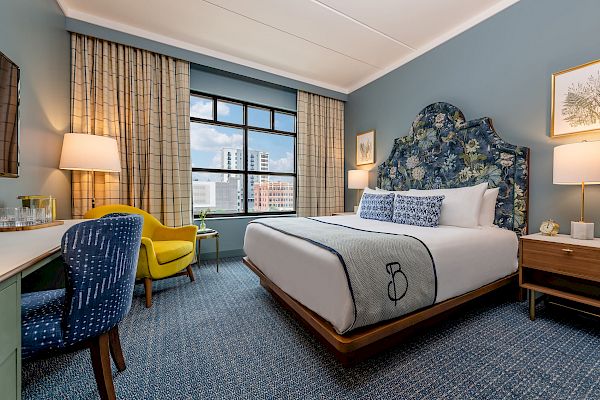 A neatly arranged hotel room features a bed with patterned headboard, a desk, a yellow chair, lamps, and a view of buildings through the window.