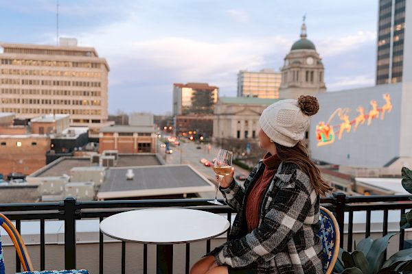 A person wearing a winter hat and holding a drink sits on a rooftop patio, overlooking a cityscape with Christmas decorations in the distance.