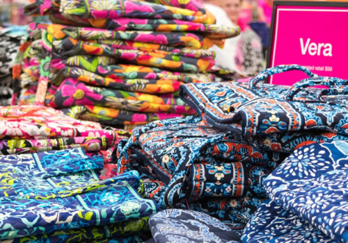 The image shows stacks of colorful, patterned bags on a display table in a store, with a pink sign labeled 