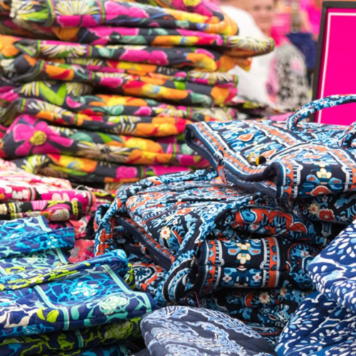 The image shows stacks of colorful, patterned bags on a display table in a store, with a pink sign labeled 