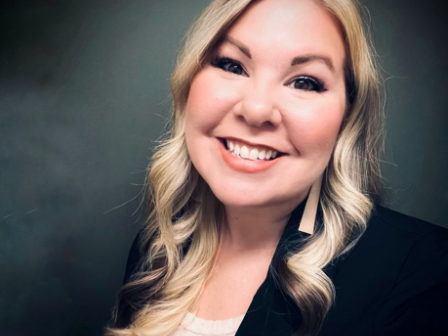 A person with long, wavy blonde hair, wearing a black blazer and smiling at the camera against a dark, neutral background.