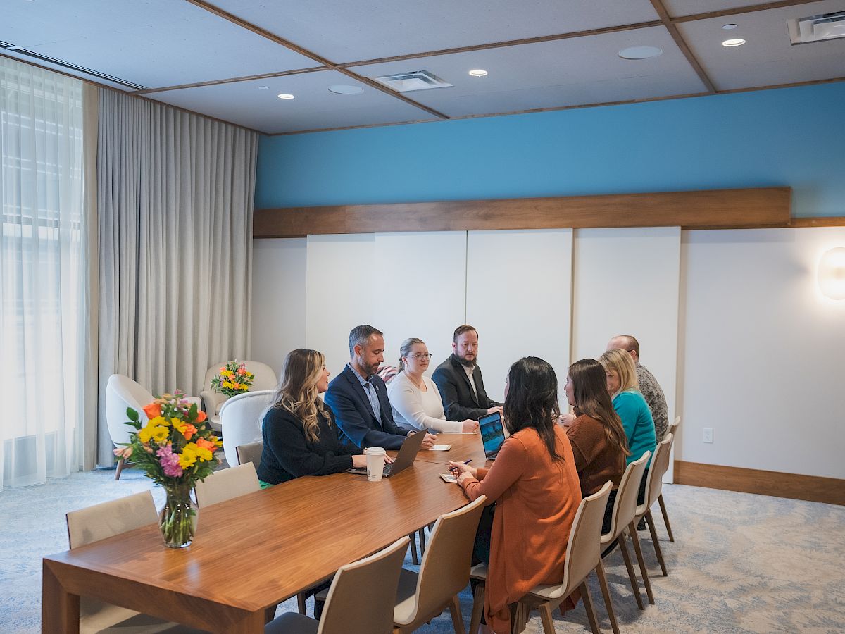 A group of people are having a meeting at a long table in a conference room with a vase of flowers. The sentence ends.