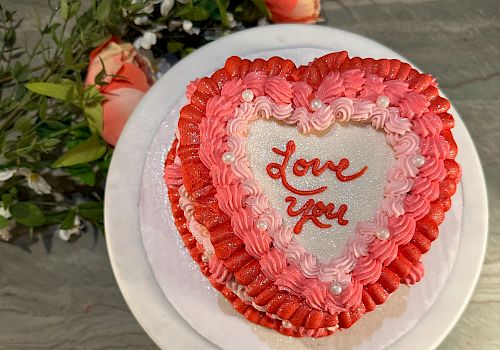 A heart-shaped cake with red and pink frosting, displaying the words 