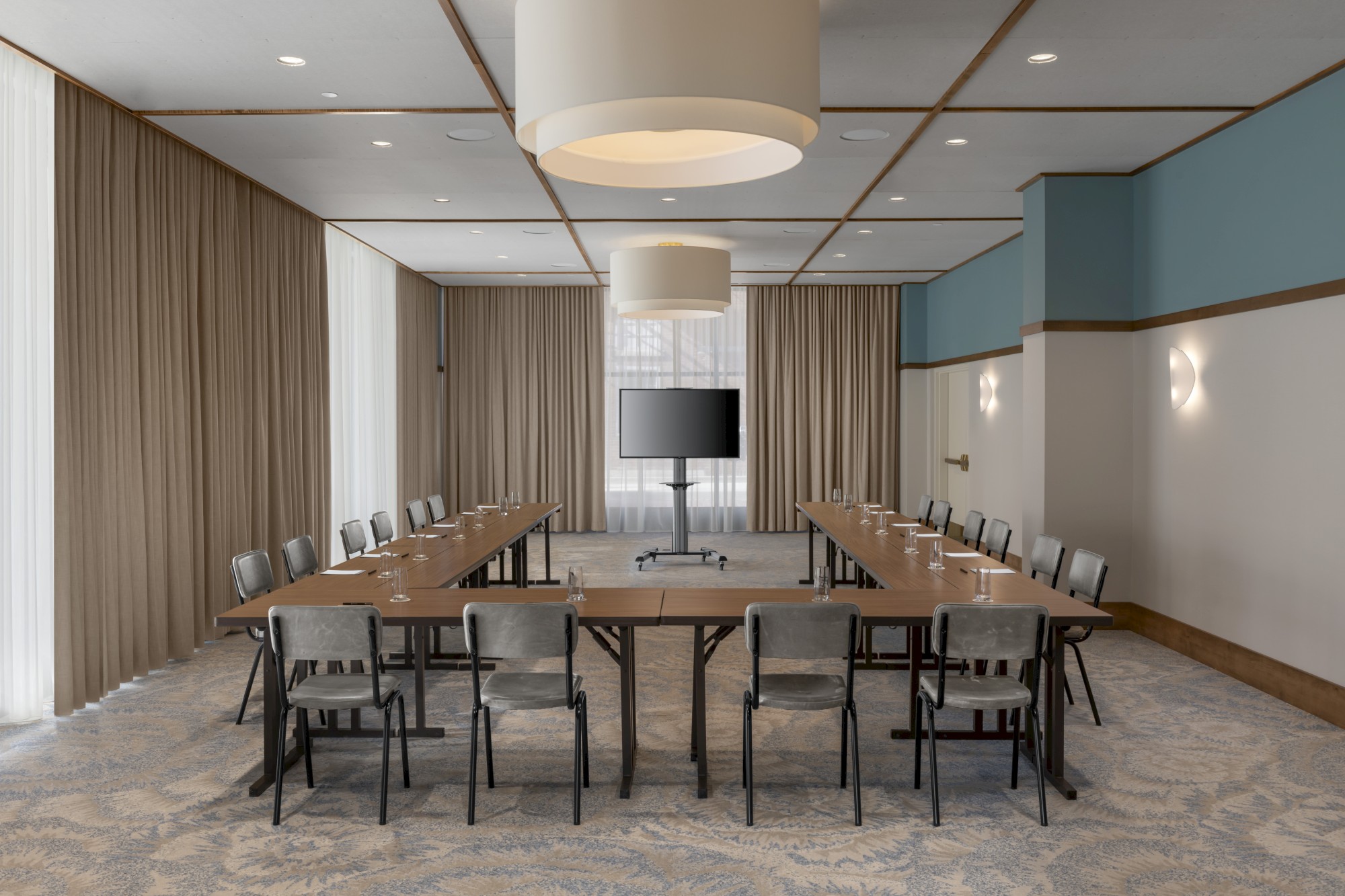 A conference room with a U-shaped table setup, chairs, a large screen, and ceiling lights, surrounded by beige curtains.