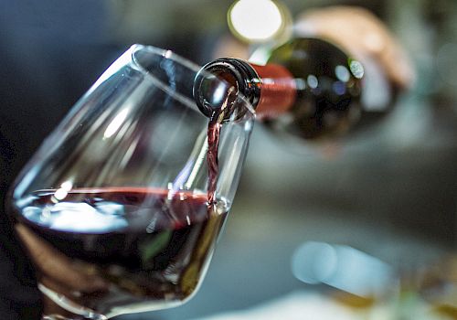 A hand pours red wine from a bottle into a glass, with a blurred background indicating a casual setting.