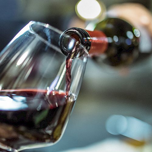 A hand pours red wine from a bottle into a glass, with a blurred background indicating a casual setting.