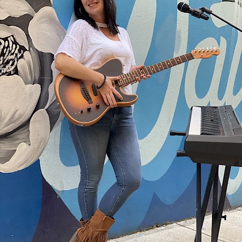 A woman stands with a guitar beside a keyboard in front of a mural, wearing jeans and fringed boots, smiling at the camera.