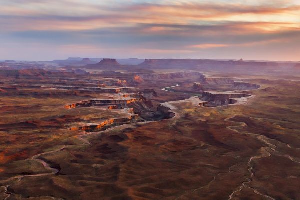The image shows a vast, arid landscape with deep canyons and winding riverbeds under a colorful sky at sunset.