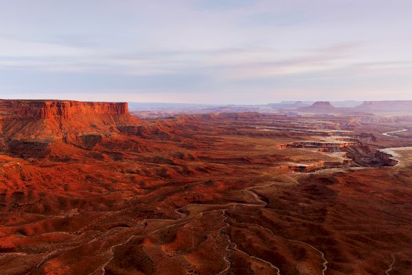 The image shows an expansive desert landscape with rocky mesas, deep canyons, and a vast open sky, captured during sunset with a warm, reddish hue.