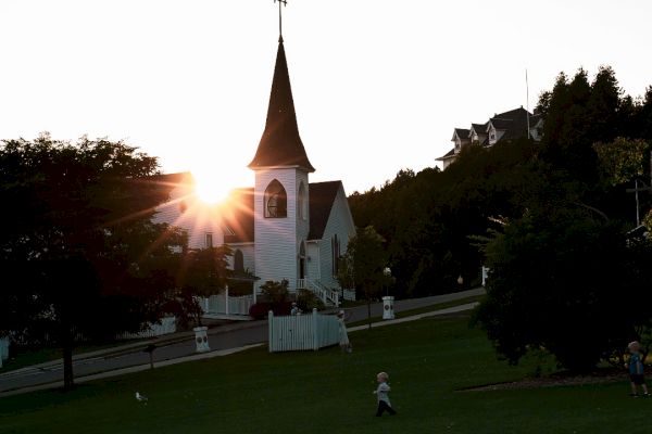 A small church with a steeple and cross is seen at sunset, surrounded by trees and grass, with people walking on a lawn in the foreground.