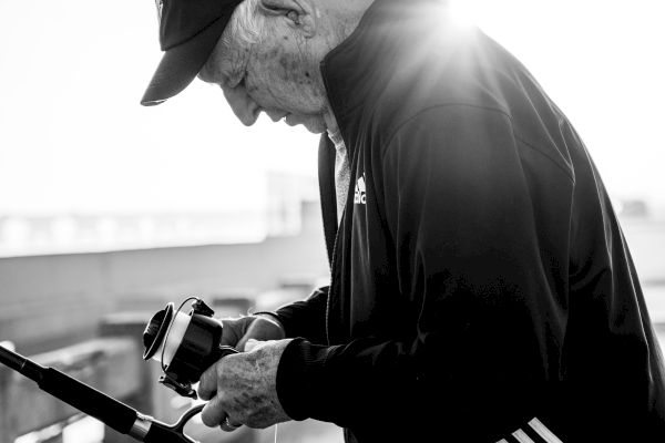The image shows an elderly man in a cap and jacket, focusing on adjusting his fishing rod, with sunlight creating a halo effect behind him.