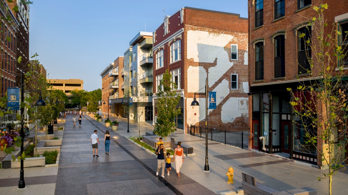 A pedestrian-friendly street features modern buildings, greenery, and people walking leisurely on a sunny day, creating a lively atmosphere.