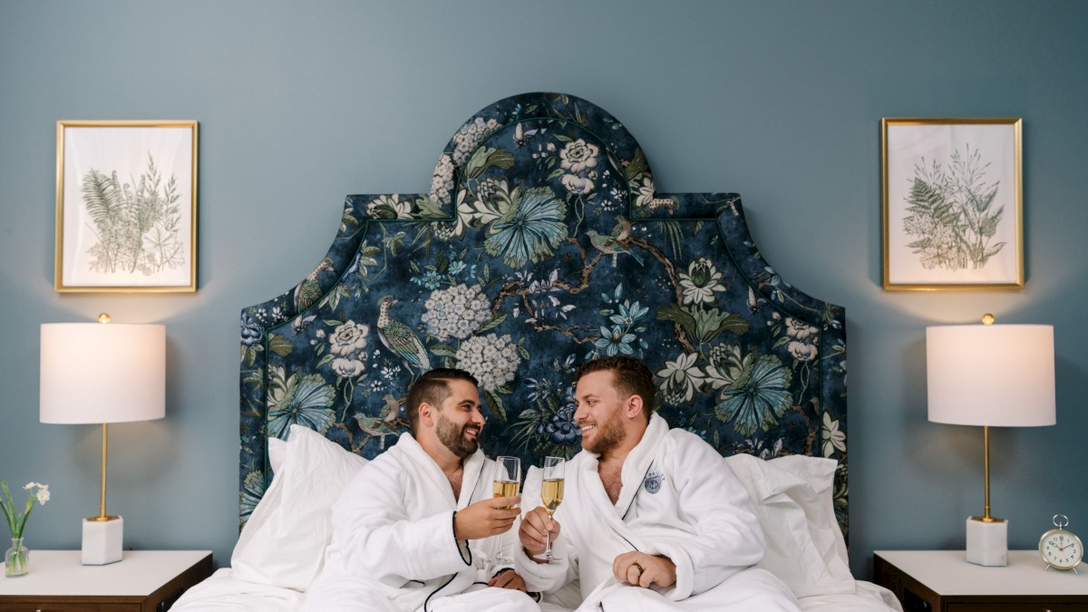 Two people in robes sit on a bed, toasting with champagne. The room has blue walls, floral headboard, matching lamps, and botanical art.