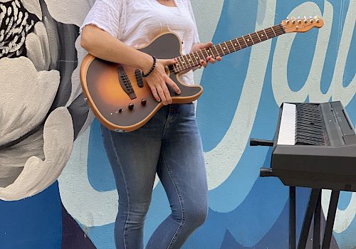 A woman holds an electric guitar, standing beside a keyboard, in front of a colorful mural featuring a flower and stylized text.