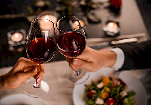 Two people are clinking wine glasses over a table with food and candles, suggesting a romantic dinner setting.