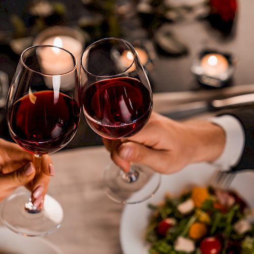 Two people are clinking wine glasses over a table with food and candles, suggesting a romantic dinner setting.