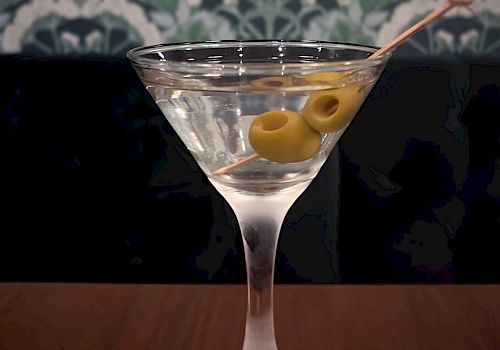 A martini glass filled with a clear cocktail garnished with two green olives on a toothpick, set against a patterned wallpaper background.