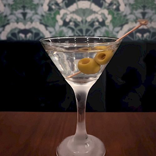 A martini glass filled with a clear cocktail garnished with two green olives on a toothpick, set against a patterned wallpaper background.
