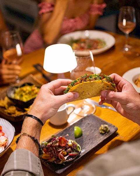A close-up of a person holding a taco at a dining table with various dishes and drinks, surrounded by others enjoying a meal.