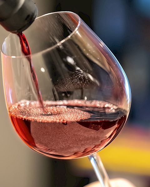 Red wine is being poured into a tilted wine glass, creating bubbles. The background is blurred, focusing attention on the pouring action.