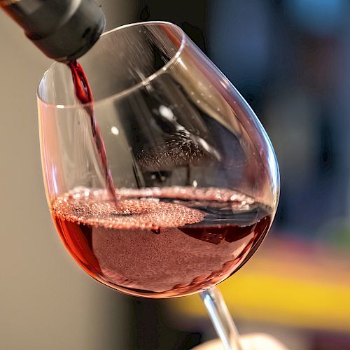 Red wine is being poured into a tilted wine glass, creating bubbles. The background is blurred, focusing attention on the pouring action.