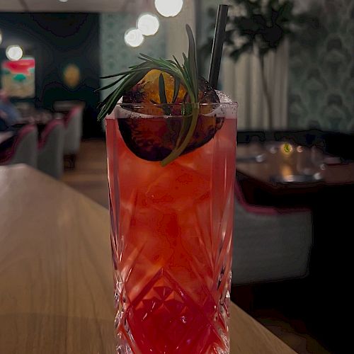 A red cocktail in a tall, patterned glass garnished with an orange slice, herbs, and a straw. The drink is placed on a wooden table.