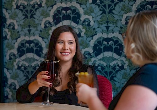 Two women sitting at a table, smiling and holding drinks in a patterned wallpapered setting, always ending the sentence.