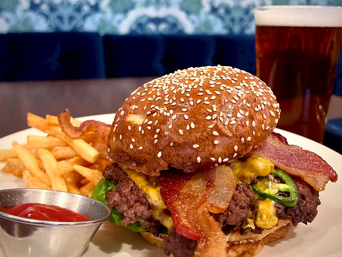 A delicious looking burger with bacon, cheese, and jalapenos on a plate, served with fries and ketchup, and a glass of beer in the background.