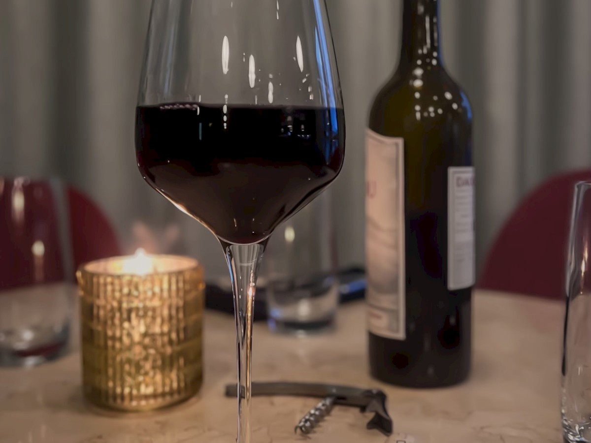 A glass of red wine, a lit candle, a wine bottle, a cork, and a corkscrew on a table. The setting looks cozy and intimate.