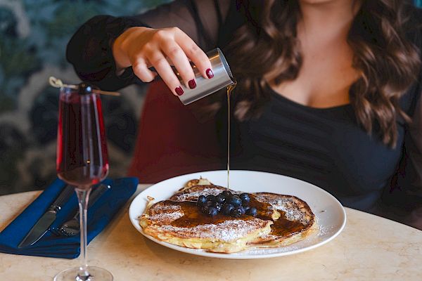 A person is pouring syrup on a pancake topped with berries, with a glass of red drink on the table.