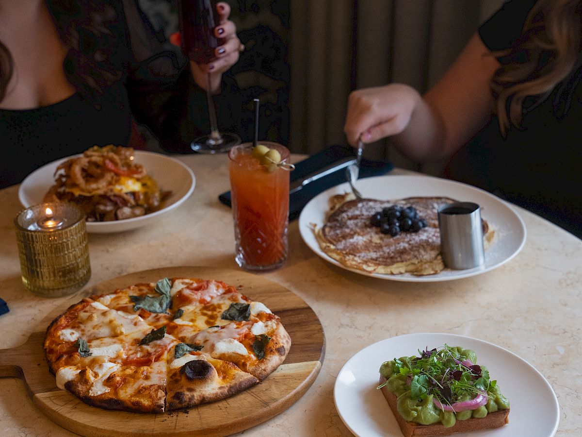 A table with dishes including pizza, pancakes, avocado toast, a candle, and two drinks with two people dining together at the table.