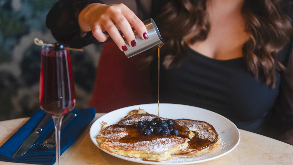 A person pouring syrup on pancakes topped with blueberries, with a glass of a red drink and a blue napkin beside the plate on the table.