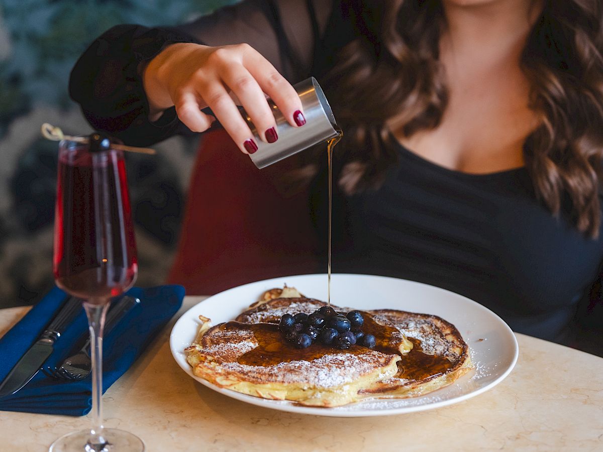 A person pouring syrup on pancakes topped with blueberries, with a glass of a red drink and a blue napkin beside the plate on the table.