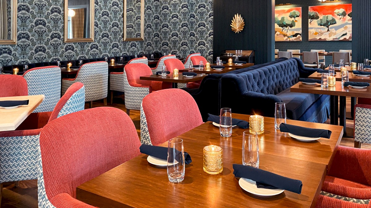 A stylish restaurant with red and blue chairs, wooden tables, patterned wallpaper, framed mirrors, paintings, and pendant lights on the ceiling.