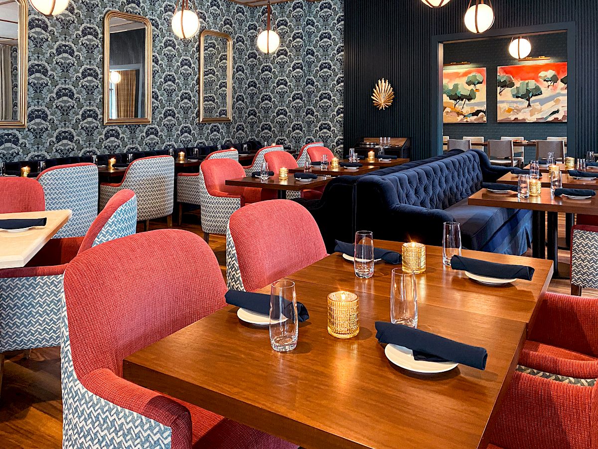 A stylish restaurant with red and blue chairs, wooden tables, patterned wallpaper, framed mirrors, paintings, and pendant lights on the ceiling.