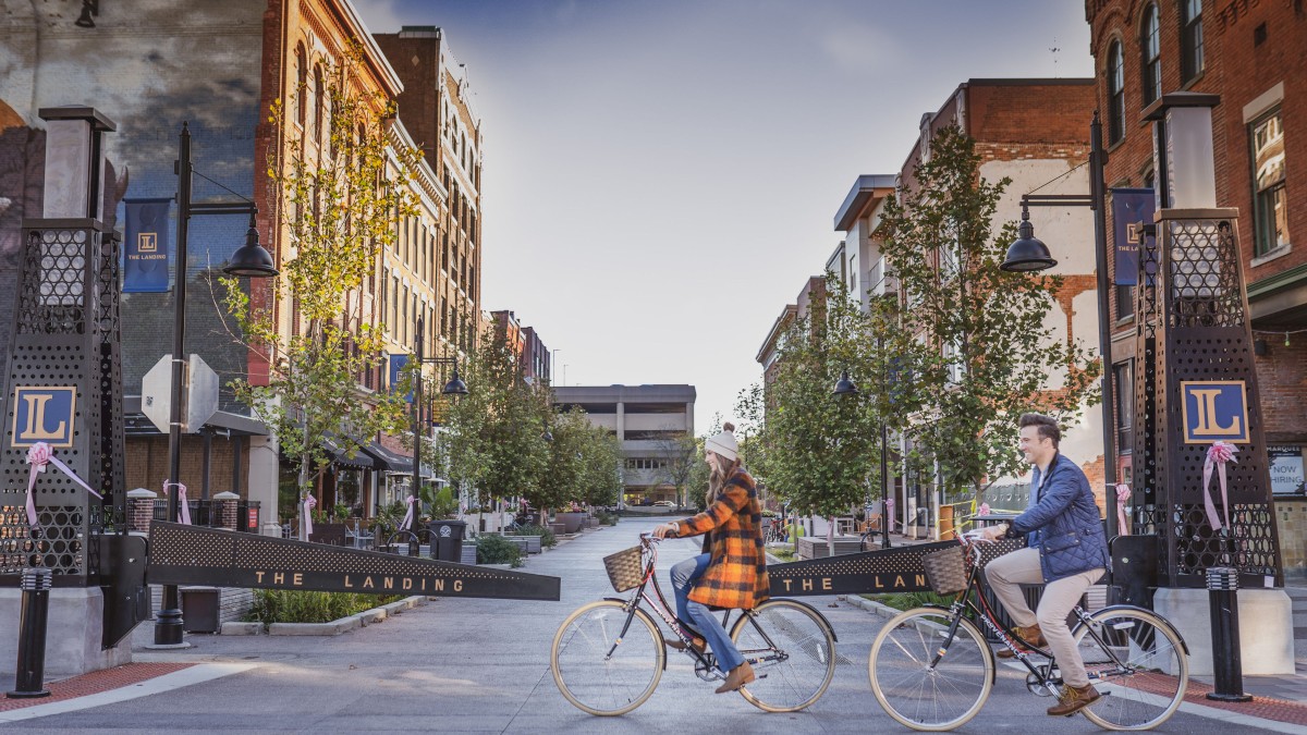 Two people ride bicycles through a gated area in a city street scene, surrounded by buildings and trees.