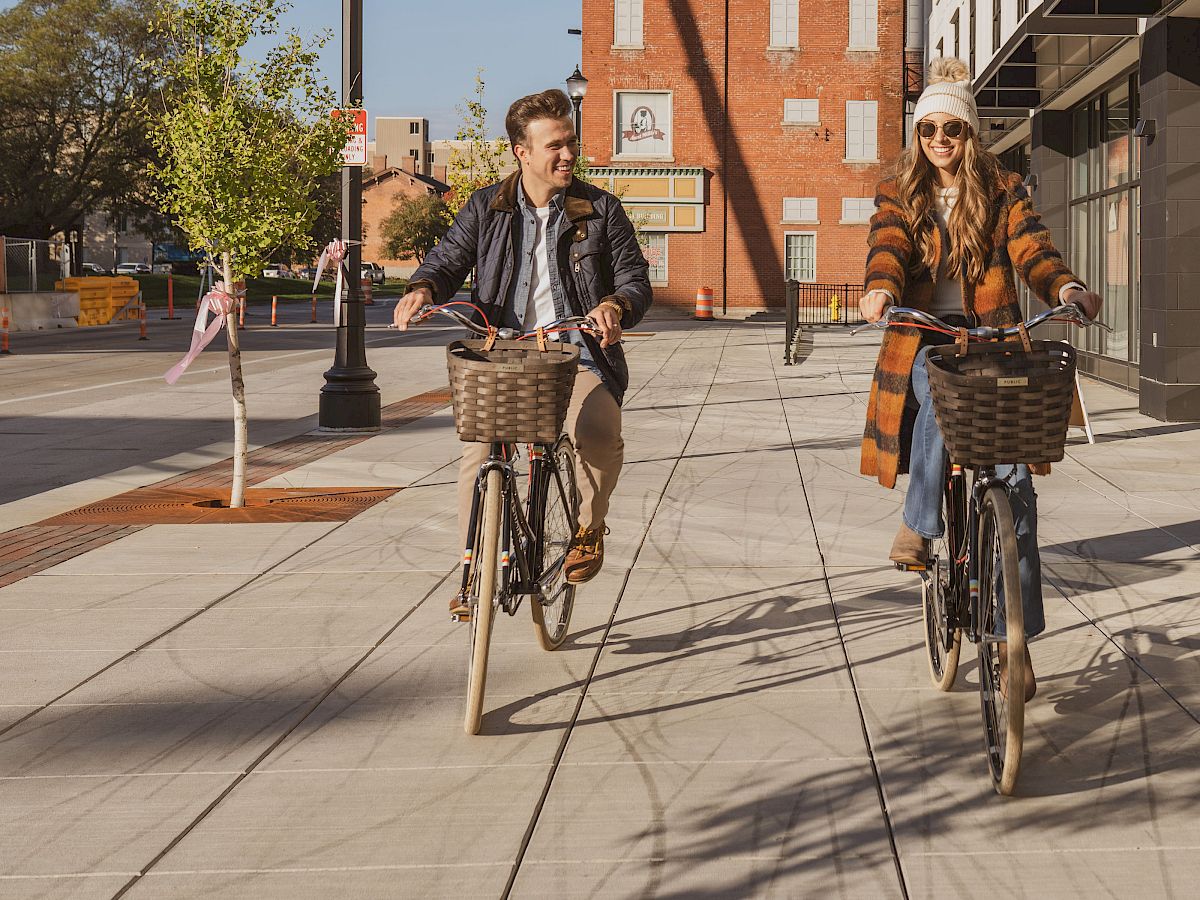 Two people are riding bicycles on a city sidewalk, smiling and enjoying a sunny day. Both have baskets on their bikes.