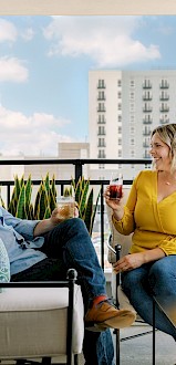 A man and woman are sitting on a balcony, smiling and having drinks. They are surrounded by plants and city buildings are visible in the background.