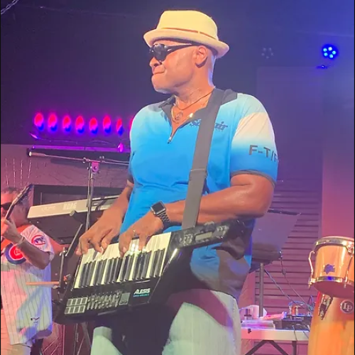 A musician plays a keytar on stage wearing a hat and sunglasses.