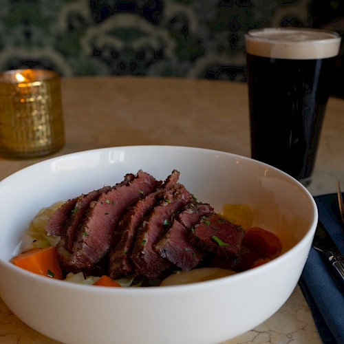 A bowl of sliced meat and vegetables, a candle, a dark beer, and cutlery on a napkin on a table.