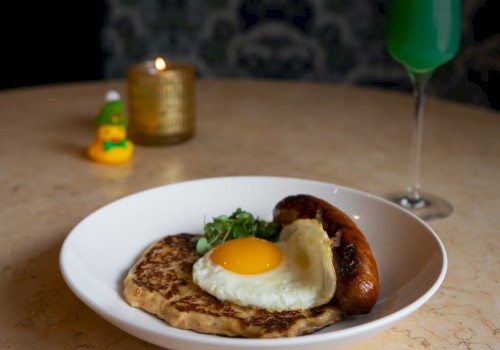 A plate with a fried egg, sausage, and pancake, placed on a table with a green drink, a candle, and a small duck figurine in the background.