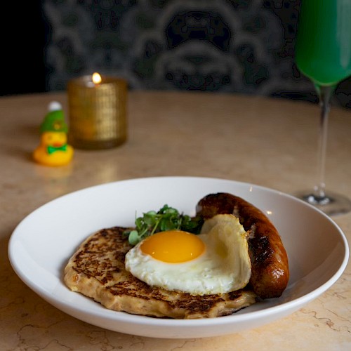 A plate with a fried egg, sausage, and pancake, placed on a table with a green drink, a candle, and a small duck figurine in the background.