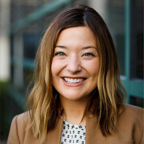 A person with shoulder-length hair is smiling, wearing a brown blazer over a patterned top, with a blurred background of what appears to be a building.