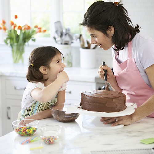 A woman and a child are decorating a chocolate cake in a bright kitchen, smiling and sharing a joyful moment.